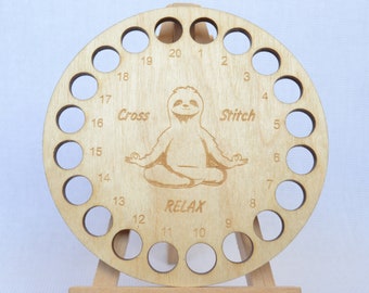 May include: A wooden thread holder with 20 holes for storing embroidery floss. The holder is round and features a sloth in a yoga pose with the words "Cross Stitch Relax" around it.