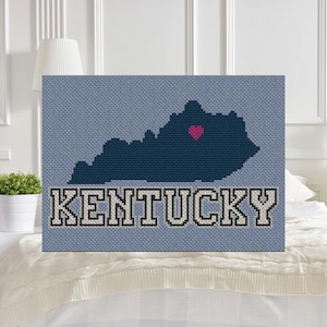 May include: A blue and white cross-stitch design of the state of Kentucky with a pink heart in the center. The word "KENTUCKY" is stitched in white letters with black outlines below the state outline.