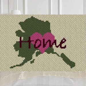 May include: A green crochet blanket with the outline of Alaska and a pink heart with the word "Home" in maroon lettering.