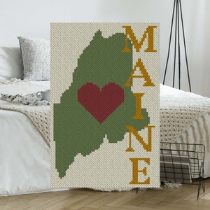 May include: A green outline of the state of Maine with a red heart in the center. The word "MAINE" is written vertically in gold letters to the right of the state outline.