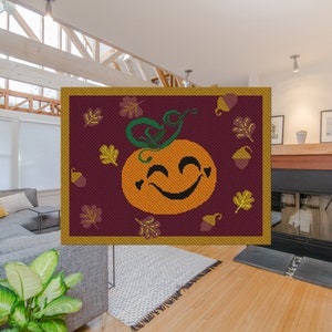 Op de afbeelding: Een vierkant wandkleed met een glimlachende pompoen en herfstbladeren in tinten oranje, groen, bruin en goud op een bordeauxrode achtergrond.