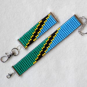 May include: Two beaded bracelets with a green, yellow, and blue color pattern, reminiscent of the Tanzanian flag. The bracelets have silver clasps and a silver chain on one of the bracelets.