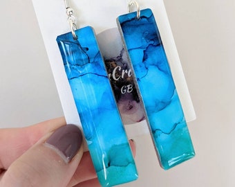 May include: A pair of blue and teal resin earrings with silver hooks. The earrings are rectangular in shape and have a marbled effect.