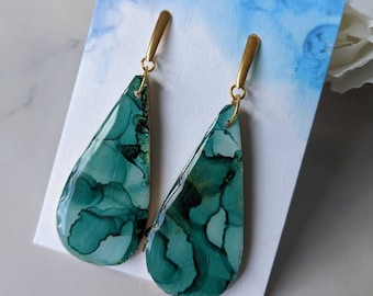 Op de afbeelding: Druppelvormige oorbellen met een marmerpatroon in teal en groen. De oorbellen hebben goudkleurige pinnen en haken. De oorbellen worden gepresenteerd op een witte kaart met een blauwe aquarel achtergrond, met witte bloemen op de achtergrond.