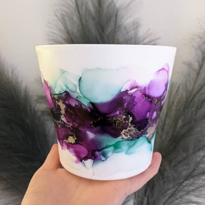 May include: White ceramic planter with an abstract design featuring purple, teal, and gold accents.