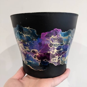 May include: Black ceramic planter with a galaxy-inspired design featuring swirls of purple, blue, and gold paint.