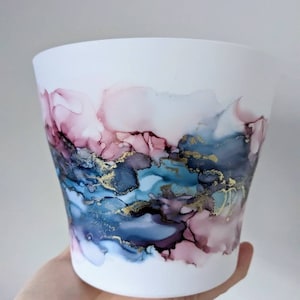 May include: White ceramic planter with a hand-painted abstract design in shades of pink, blue, and gold. The design features swirling, marbled patterns.
