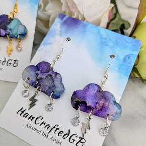 May include: Purple and blue cloud-shaped earrings with silver lightning bolt and crystal accents. The earrings are on a white card with the text "HandCraftedGB Alcohol Ink Artist".