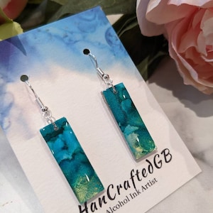 May include: A pair of teal and gold alcohol ink earrings with silver hooks. The earrings are rectangular in shape and have a marbled effect. The card they are displayed on reads "HandcraftedGB Alcohol Ink Artist".