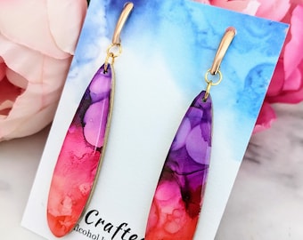 Op de afbeelding: Een paar oorhangers met een traanvormige hanger van hars. De hanger is een gradiënt van roze, rode en paarse alcoholinkt. De oorbellen hebben goudkleurige haakjes. De kaart staat "Crafted Alcohol Ink Artist".