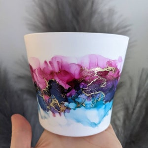 May include: White ceramic planter featuring an abstract design in shades of pink, blue and gold. The design incorporates swirls and streaks of colour, creating a unique and artistic look.
