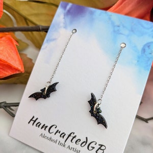 May include: A pair of black bat dangle earrings with silver chains. The earrings are on a white card with the text "HanCraftedGB Alcohol Ink Artist".