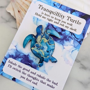 May include: A blue and gold resin turtle figurine with the text "Tranquillity Turtle is here to keep you well. Hold me tight and rub my shell. Inhale the good and exhale the bad. I'll switch the thoughts that make you feel sad."