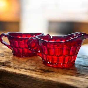 May include: Two pieces of ruby red glass tableware. One is a small creamer with a spout and handle, and the other is a small cup with a handle. Both pieces have a textured, geometric pattern.