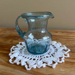 May include: A small, clear blue glass pitcher with a handle. The pitcher is sitting on a white crocheted doily.