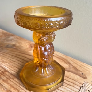 Puede incluir: Portavelas de vidrio ámbar con un diseño decorativo de querubines y motivos florales. El portavelas tiene una parte superior redonda y una base hexagonal.