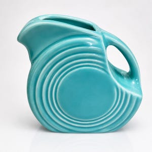 Puede incluir: Una jarra de cerámica de color turquesa con un acabado brillante y una serie de anillos concéntricos en relieve en el lateral. El recipiente tiene un asa curva, un pico vertedor ancho y una forma redondeada con un color uniforme y liso.