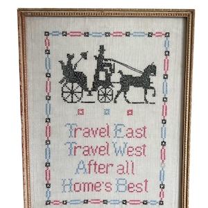 May include: Framed cross-stitch embroidery with a black silhouette of a horse-drawn carriage and the text "Travel East Travel West After all Home's Best". The embroidery is on a white linen fabric with a blue and pink border.