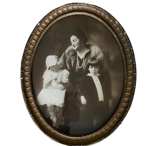 Puede incluir: Una fotografía en blanco y negro de una mujer y dos niños en un marco ovalado de oro. La mujer lleva un abrigo de piel y un collar de perlas. Los niños llevan ambos vestidos. La foto es de estilo vintage.