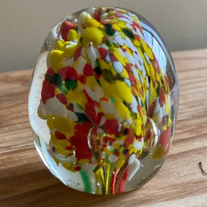 Op de afbeelding: Een transparante glazen brievengewicht met een werveling van rood, geel, wit en groen glas erin.