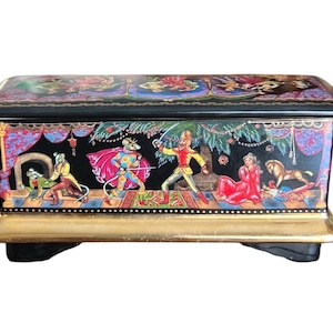 Puede incluir: Una caja decorativa rectangular negra con base y patas doradas. La caja presenta una colorida ilustración de una escena de cuento de hadas con figuras disfrazadas. La tapa está decorada con motivos florales y ornamentales.