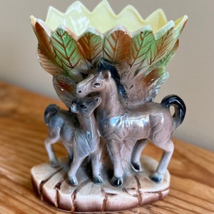 May include: A ceramic vase shaped like a tree with two horses standing beneath it. The vase is decorated with brown, green, and yellow paint. The horses are brown and gray.