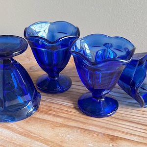 Puede incluir: Cuatro platos de postre de vidrio azul cobalto. Tres de los platos tienen una forma tradicional de sundae con un borde acanalado. El cuarto plato es de estilo pedestal con un borde acanalado.