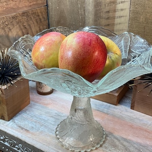 May include: A clear, glass pedestal fruit bowl filled with three red and yellow apples. The bowl has a ruffled edge and sits on a decorative base. The background is a wooden wall.