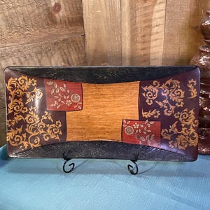 May include: A rectangular decorative tray featuring a central light wood panel flanked by two dark red sections with gold floral scrollwork and two black textured ends. The tray is displayed on a black metal stand, showcasing a multi-toned finish.