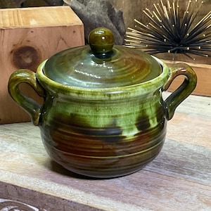 May include: A round ceramic sugar bowl with two side handles and a matching lid featuring a small knob. The vessel has a glossy glaze with a gradient of deep forest green at the top transitioning into earthy brown tones at the base.