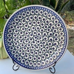 May include: A round ceramic plate featuring a dense, repeating pattern of small blue and dark purple flowers on a cream-colored background, displayed on a black metal stand. The design covers the entire surface in a swirling, circular arrangement.