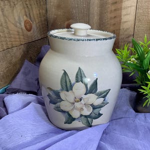 May include: A cream-colored ceramic canister with a lid, featuring a hand-painted magnolia flower design with green leaves on the front. The rim of the canister and the lid edge are decorated with a dark blue speckled pattern on the ceramic surface.