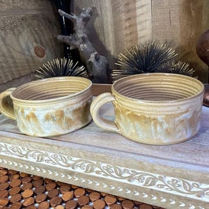 May include: Two ceramic soup mugs with a wide, low-profile design and a single handle. The mugs feature a two-tone glaze with a creamy off-white top section that transitions into a warm, mottled tan and brown base with visible horizontal ridges.