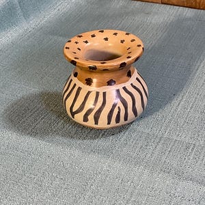 May include: A small, round, tan ceramic vase with a wide, flat rim. The rim is decorated with small, dark brown, star-like markings. The body of the vase features vertical, dark brown, wavy stripes that extend from the base to the shoulder.
