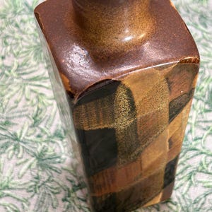 May include: A square-shaped ceramic vase with a narrow neck. The vase features a patchwork design in shades of brown, black, and tan. The top and neck are a solid brown color. The vase is likely for decorative purposes.
