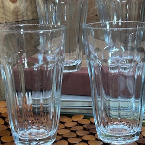 May include: A collection of clear, ribbed drinking glasses. The glasses are made of transparent glass and have a classic, simple design. The glasses are arranged on a wooden surface, suggesting a rustic or vintage aesthetic. The glasses are suitable for water, juice, or other beverages.