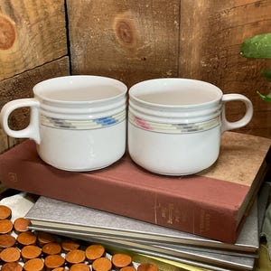 May include: Two white ceramic mugs with a horizontal band of pastel colors. The mugs have a rounded shape with a handle and are sitting on top of a stack of books. The background is a wooden surface.