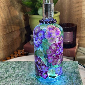May include: A decorative glass bottle with a floral design in shades of purple and blue. The bottle is illuminated with blue lights and has a mirrored top. The bottle is adorned with dark purple beads.