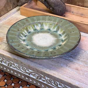 May include: A round ceramic bowl with a wide, fluted rim featuring a mottled olive green glaze. The center of the bowl transitions into a smooth, creamy white basin with subtle blue-green speckling and reactive glaze patterns throughout the surface.
