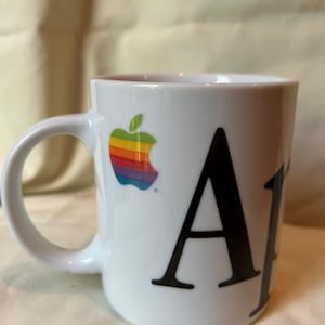 May include: White ceramic mug featuring the classic Apple logo in rainbow colors and the letter "A" in black. The mug has a curved handle and a simple, clean design, perfect for coffee or tea.