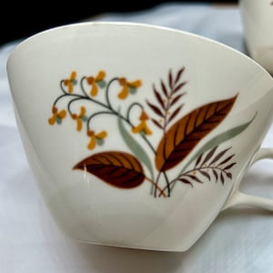 May include: Cream-colored teacup with a floral design featuring brown, yellow, and green leaves and flowers. The cup has a curved shape and a small handle. A second cup is partially visible in the background.