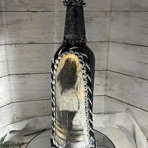 May include: A black glass bottle decorated with silver and black beads, a black and white image of a woman, and a silver and black beaded cap. The bottle is sitting on a silver mirror.