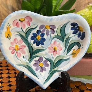 May include: Heart-shaped ceramic dish with a hand-painted floral design. The dish features pink, purple, blue, and yellow flowers with green leaves against a white background. The rim is light blue. The dish is displayed on a black stand.