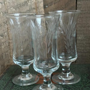 May include: Three clear, stemmed glasses with etched floral designs. The glasses have a tulip shape and sit on a round base. The background is a weathered wooden surface.