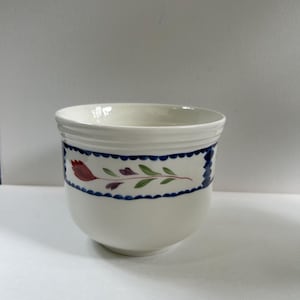 May include: A white ceramic cup with a floral design. The cup has a blue scalloped border with red and purple flowers and green leaves. The cup is round with a slightly flared rim.