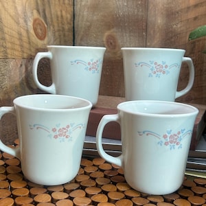 May include: Four white ceramic mugs with a floral design in pink and blue. Each mug has a curved handle and a simple, elegant shape. The mugs are arranged on a wooden surface.