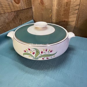 May include: A round white ceramic casserole dish with a matching lid featuring a teal center band and a silver rim. The body of the dish is decorated with a floral pattern of red and yellow tulips with green leaves and a silver trim on the handles.