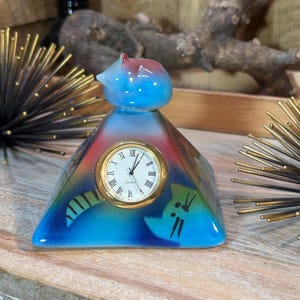 May include: A ceramic cat-shaped clock with a quartz movement. The clock has a blue, red, and green color scheme. The clock face is gold with Roman numerals. The cat has a blue head and a red top.