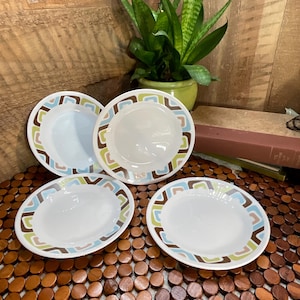 May include: Four white ceramic plates with a retro design. Each plate has a rim decorated with brown, green, and blue geometric shapes. The plates are arranged on a wooden surface, with a plant in the background.