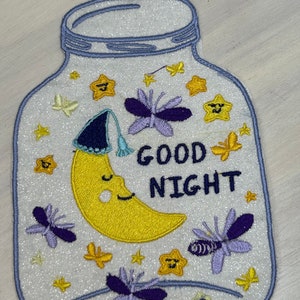 May include: A white, glittery, embroidered patch shaped like a jar with a blue lid. Inside the jar is a yellow crescent moon with a blue sleeping cap, purple butterflies, and yellow stars. The text "GOOD NIGHT" is embroidered in black.